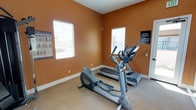 This single-floor fitness center offers a focused workout environment designed for cardio training. The room is finished with warm terracotta-orange walls complemented by white baseboards, creating an energetic yet welcoming atmosphere. Natural light filters in through multiple windows fitted with horizontal blinds, brightening the space and providing views of the surrounding exterior. The flooring features a neutral, low-pile carpet that provides a practical surface underfoot. The room is equipped with key cardio machines, including an elliptical trainer and a treadmill, positioned to make efficient use of the available floor space. An exercise instruction poster is mounted on the wall, offering visual guidance for users. Informational signage is also posted, indicating facility rules and operating hours, including alarm activation after closing time. The overall layout is straightforward and functional, prioritizing accessibility to equipment. The space is well-maintained with clean walls and tidy equipment arrangement, making it a practical option for residents or users seeking a convenient, no-frills cardio workout facility.