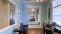 This single-story space presents a refined and carefully curated interior defined by its soothing blue walls and rich hardwood flooring. The main area serves as a welcoming reception or lounge space, furnished with a plush navy blue accent chair and a sleek round side table. Framed botanical artwork adorns the walls, adding a touch of nature-inspired elegance to the cool color palette. White crown molding provides a crisp architectural contrast against the blue walls, lending a classic and polished finish throughout.

A wide, white-trimmed archway connects the front area to an inner room, drawing the eye toward a striking chandelier with an artistic, multi-arm design that serves as a clear focal point. The inner room features dark cabinetry, large mirrors, and additional chandelier lighting, suggesting a professional service environment such as a salon or medical/aesthetic suite. Natural light filters in through multiple windows, enhancing the bright and airy atmosphere. The cohesive use of blue tones, white trim, and warm wood flooring throughout creates a consistent, sophisticated aesthetic that feels both professional and inviting.
