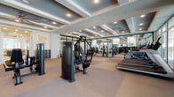 Located at 6087 Fruitville Rd in Sarasota, Florida, this single-story fitness and wellness facility offers a comprehensive range of amenities designed to support both athletic performance and recovery. The space is thoughtfully organized into distinct zones, each serving a specific purpose within an overall clean, modern aesthetic defined by white walls, light wood-look flooring, and contemporary finishes.

The centerpiece of the facility is a generously sized fitness floor equipped with an extensive array of strength machines, cable systems, free weights, and cardio equipment including treadmills and ellipticals. Coffered ceilings with ceiling fans add architectural interest overhead, while expansive windows along one wall flood the space with natural light and offer outdoor views. Full-length mirrors amplify the sense of openness throughout the gym area.

Adjacent to the fitness floor, a stylish lounge area provides a welcoming transition space, furnished with teal accent chairs, a neutral bench seat, and mirrored wall panels. A glass-enclosed golf simulator room adds a distinctive recreational element, featuring a high-definition projection screen and synthetic turf hitting mat.

Dedicated recovery or treatment rooms offer a quieter, more intimate setting, furnished with reclining chairs and soft lighting, suitable for wellness treatments or relaxation sessions.