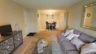This single-floor residence presents an open-concept layout that seamlessly connects the living and dining areas, creating a cohesive and functional flow. The living area is anchored by a gray upholstered sofa accented with decorative pillows, complemented by a wood-finished coffee table and a patterned area rug that adds texture to the carpeted flooring. Warm, neutral tones on the walls contribute to a calm and inviting ambiance throughout the space.

The dining area is positioned just beyond the living room, defined by a pendant light fixture that hangs above a dark-finished dining table surrounded by matching chairs. Framed wall art is displayed behind the dining set, adding a decorative focal point to the space. Carpeting continues seamlessly from the living area into the dining zone, reinforcing the unified aesthetic.

The entry foyer is visible to the left, featuring a white door with a doormat, providing a clear and practical transition point into the home. Overhead, recessed ventilation and subtle architectural details give the space a clean, finished appearance. Overall, the layout is well-suited for everyday living and casual entertaining.
