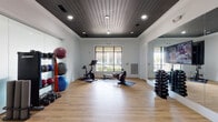 This single-floor fitness room offers a comprehensive workout environment designed to support a wide range of exercise routines. The space is defined by its striking dark wood plank ceiling accented with recessed lighting, which contrasts elegantly with the bright white walls and clean crown molding to create a polished, modern aesthetic. Warm-toned hardwood flooring runs throughout, providing a durable yet attractive surface underfoot.

The room is equipped with a variety of cardio and strength-training equipment, including a stationary spin bike and a rowing machine, both positioned on protective floor mats near large windows that allow ample natural light and views to the outdoors. A full-length wall mirror on one side enhances the sense of space and supports proper form during workouts.

Along the walls, organized storage racks hold a comprehensive collection of stability balls in assorted sizes, medicine balls, resistance bands, kettlebells, and a full dumbbell rack ranging across multiple weight increments. Adjustable weight benches provide additional versatility for strength training. A frosted glass entryway adds a refined, modern touch to the entrance. The overall ambiance is clean, functional, and thoughtfully designed to support a thorough fitness experience.