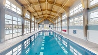 Located at 1111 W Bay Ave in Barnegat, NJ, this impressive indoor aquatic facility is a single-floor structure spanning a generously sized footprint. The space is defined by its dramatic vaulted timber-frame ceiling, where exposed wooden arched beams create a warm, barn-inspired architectural aesthetic that rises high overhead, lending a grand sense of volume and openness to the interior.

The facility's primary feature is a full-length lap pool with clearly marked lane lines, ideal for recreational swimming and fitness. Adjacent to the main pool is a separate, smaller therapy or spa pool equipped with an accessibility lift, ensuring inclusive use for all visitors. The pool deck is finished in large-format stone tiles, providing a clean and safe surround.

Natural light floods the space through an expansive wall of floor-to-ceiling glass panel garage-style doors along one side, as well as large multi-pane windows that wrap the upper portions of the walls. These openings offer views of the surrounding outdoor grounds and greenery, blurring the line between indoor and outdoor environments.

The interior walls are painted in soft blue and white tones, complementing the aqua hues of the pool water. Poolside amenities include white lounge chairs, safety equipment, first aid stations, and clearly posted pool rules, reflecting a well-maintained and safety-conscious facility.