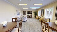 Located at 540 Deauville Dr in Monroeville, PA, this single-floor community amenity facility offers a well-rounded set of shared spaces designed for comfort and active living. The entrance welcomes visitors into a bright, warmly decorated lounge area with warm yellow tones, patterned carpet, recessed and skylight-style ceiling fixtures, crown molding, and comfortable seating arrangements centered around a wall-mounted TV. Large windows fitted with blinds allow natural light to fill the space. Adjacent to the lounge, the dining room provides a relaxed gathering area with multiple round wooden dining tables and chairs, additional natural light from exterior-facing windows, and direct access to connecting rooms. The fitness center is divided into two dedicated exercise rooms, both clad in rich wood-paneled walls and ceilings that lend a warm, cabin-like ambiance. The first room houses cardio equipment including treadmills, an elliptical, and a stationary bike, while the second offers a comprehensive strength-training setup with cable machines, free weights, benches, and wall-mounted mirrors. Outside, an expansive pool area features lounge chairs on a paved deck, along with fenced tennis courts and open green surroundings, completing this versatile community space.