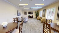 Located at 540 Deauville Dr in Monroeville, PA, this single-floor community amenity facility offers a well-rounded set of shared spaces designed for comfort and active living. The entrance welcomes visitors into a bright, warmly decorated lounge area with warm yellow tones, patterned carpet, recessed and skylight-style ceiling fixtures, crown molding, and comfortable seating arrangements centered around a wall-mounted TV. Large windows fitted with blinds allow natural light to fill the space. Adjacent to the lounge, the dining room provides a relaxed gathering area with multiple round wooden dining tables and chairs, additional natural light from exterior-facing windows, and direct access to connecting rooms. The fitness center is divided into two dedicated exercise rooms, both clad in rich wood-paneled walls and ceilings that lend a warm, cabin-like ambiance. The first room houses cardio equipment including treadmills, an elliptical, and a stationary bike, while the second offers a comprehensive strength-training setup with cable machines, free weights, benches, and wall-mounted mirrors. Outside, an expansive pool area features lounge chairs on a paved deck, along with fenced tennis courts and open green surroundings, completing this versatile community space.