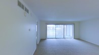 This single-floor space features an open living area designed with simplicity and functionality in mind. The room is finished with wall-to-wall carpeting in a neutral tone, providing a soft and comfortable foundation throughout. Crisp white walls contribute to a clean, bright aesthetic that feels open and airy.

A prominent feature of the living area is the large sliding glass door dressed with vertical blinds, which serves as the primary source of natural light. The door opens to an outdoor view, allowing ample daylight to fill the interior and creating a seamless connection between the indoor and outdoor environments. A secondary window near the entry door further enhances the natural light in the space.

Overhead, a ceiling-mounted HVAC vent ensures climate control throughout the room, while a smoke detector is also visible, reflecting standard safety provisions. A wall-mounted thermostat is conveniently positioned for easy access. The overall layout is open and unobstructed, offering flexibility for a variety of furniture arrangements. The combination of neutral finishes, natural light, and practical features makes this a functional and comfortable living space.