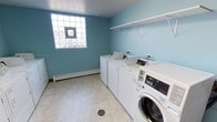 This single-floor laundry facility is designed with practicality and efficiency in mind. The room is finished with light teal walls that give the space a clean, refreshing atmosphere, complemented by durable, easy-to-maintain tile flooring. Natural light filters into the room through a distinctive glass block window, adding brightness while maintaining privacy. A baseboard heating unit runs along the exterior wall, ensuring comfort during colder months. The room is equipped with multiple top-loading washing machines and at least one front-loading washer-dryer unit, providing ample capacity for shared use. The machines are modern commercial-grade appliances with digital controls for user-friendly operation. A sturdy wall-mounted shelf with brackets spans one wall, offering convenient space for laundry supplies and folded items. A floor drain is centrally located, ensuring easy cleanup and maintenance. The layout is straightforward and spacious, allowing multiple users to move comfortably throughout the room. Overall, this laundry facility is a well-appointed, functional space built to serve the practical needs of its users efficiently.