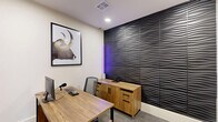 Located at 7303 S Peek Rd in Richmond, TX, this micro office is a thoughtfully designed single-room workspace that maximizes both style and functionality within a compact footprint. The office makes a strong visual impression with a dramatic floor-to-ceiling 3D wave-textured accent wall in dark charcoal, which adds depth and a contemporary edge to the interior. A subtle purple accent light nestled beside the wall introduces a modern, ambient glow that complements the room's overall aesthetic. The remaining walls are finished in a clean, neutral white, keeping the space feeling bright and open. Warm wood-toned furnishings anchor the room, including a sleek L-shaped desk with a metal frame and a matching credenza with cabinet storage — both providing ample workspace and organization. Gray carpet tiles cover the floor, adding a professional tone underfoot. Recessed ceiling lighting ensures even, focused illumination throughout. A bold framed artwork featuring a longhorn bull adds a distinctive decorative touch to the wall above the desk. The office is accessed through a black-framed glass-panel door, and a posted set of micro office rules near the entrance ensures a structured, professional environment for all users.