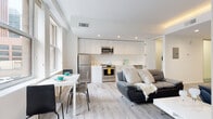 Located at 600 E Superior Ave in the heart of Cleveland, Ohio, this modern single-floor unit offers a thoughtfully designed open-concept layout that seamlessly integrates the living, dining, and kitchen areas. Light hardwood-style flooring runs throughout, complementing the bright white walls and recessed lighting that create a clean, contemporary aesthetic.

The living area is generously proportioned, offering city views through large windows that flood the space with natural light. The kitchen is fully equipped with stainless steel appliances — including a refrigerator, range, and built-in microwave — paired with sleek white cabinetry that extends to the ceiling, maximizing storage. White countertops and a modern sink complete the functional yet stylish cooking space.

A dedicated dining area adjacent to the kitchen provides a comfortable setting for meals. The bedroom is a well-appointed retreat featuring hardwood-style flooring, ample space for furnishings, and a dresser with mirror. The bathroom is finished in an all-white palette with a soaking tub and shower combination, a wall-mounted toilet, a floating vanity sink, and a medicine cabinet mirror, delivering a sleek, minimalist feel. Overall, the unit presents a polished urban living experience in a prime Cleveland location.