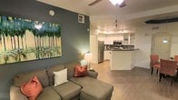 This single-floor space showcases an open-concept design that seamlessly connects the living area and kitchen, creating a cohesive and functional layout. The living area features warm hardwood-style flooring, a ceiling fan overhead, and a comfortable sectional sofa accented with decorative pillows. A bold teal-painted accent wall serves as a focal point, complemented by a large nature-inspired canvas artwork that adds color and visual interest to the space. A floor lamp provides warm ambient lighting alongside the natural light filtering into the room.

The kitchen is positioned at the rear of the open layout and is outfitted with stainless steel appliances, including a refrigerator, microwave, and range. White cabinetry with open shelving offers both storage and display space, while a dark tile backsplash adds a contemporary contrast. A peninsula-style counter provides additional workspace and serves as a natural divider between the kitchen and living areas. A pendant light hangs above, contributing to the kitchen's functional yet stylish atmosphere. Overall, the space presents a clean, modern aesthetic with a warm and inviting ambiance.