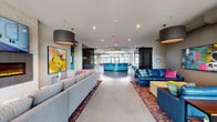 Luxor's community clubhouse is a thoughtfully designed, single-floor social hub that seamlessly blends modern aesthetics with inviting, functional spaces. The expansive main lobby greets visitors with polished concrete floors, large-scale colorful artwork, and plush seating areas anchored by richly patterned rugs. Dramatic drum pendant lights and recessed lighting illuminate the open layout, while perforated black architectural panels serve as stylish room dividers throughout.

At the heart of the space sits a striking curved bar with vibrant LED-lit panels and sleek bar stools, functioning as a social centerpiece. Large floor-to-ceiling windows flood the area with natural light and offer views of the surrounding exterior. A dedicated game room features a foosball table, a bar counter with seating, and large-format display screens for entertainment.

A cozy lounge area, labeled "Mingle," offers a warm fireplace with a mounted TV above, complemented by bold yellow accent chairs and vibrant abstract art. Additional vignettes throughout bear playful typographic wall decals such as "Imbibe," "Pour," and "Love. Life. Here." reinforcing the space's lively, community-forward character. The overall ambiance is energetic yet comfortable, designed to encourage social interaction and a sense of belonging.