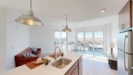 This contemporary single-floor residence showcases a thoughtfully designed open-concept layout that seamlessly connects the kitchen and living areas. The kitchen is a standout feature, equipped with rich dark wood cabinetry, stainless steel appliances including a refrigerator, range, and microwave, and a generous island with a white countertop and undermount sink. Industrial-style pendant lights hang above the island, adding a modern aesthetic touch. Light wood flooring runs throughout the main living areas, contributing to a bright and airy atmosphere. Large floor-to-ceiling sliding glass doors and expansive windows in both the living room and bedroom flood the space with natural light while offering sweeping views of the surrounding urban landscape. The living area opens directly to a private balcony, further enhancing the connection to the outdoors. A dedicated laundry closet with a stacked washer and dryer unit adds everyday convenience. The bedroom features plush carpeting and its own access to an outdoor space with city views. The bathroom is finished with large-format light tile flooring and warm wood cabinetry, maintaining the clean, modern palette found throughout. The overall ambiance is bright, contemporary, and well-appointed.
