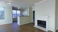 This single-floor space offers an open-concept layout that seamlessly connects the living area and kitchen. The living room is anchored by a classic white-mantled fireplace with a dark interior, adding both a functional and decorative focal point to the room. Crown molding runs along the ceiling throughout, lending a refined architectural detail to the otherwise clean, neutral aesthetic.

The kitchen is defined by a peninsula with a dark countertop, providing a natural divide between the cooking and living spaces while maintaining an open feel. White upper cabinetry and a built-in microwave are visible, reflecting a functional and straightforward design. A ceiling fan with track lighting overhead ensures the space is well-lit.

Large windows along one wall allow generous natural light to fill the interior, highlighting the rich, dark wood-style flooring that extends throughout. The neutral warm-beige wall tones complement the flooring and create a cohesive, inviting ambiance. A doorway visible in the background leads to an additional room, suggesting further usable space within the unit.