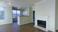 This single-floor space offers an open-concept layout that seamlessly connects the living area and kitchen. The living room is anchored by a classic white-mantled fireplace with a dark interior, adding both a functional and decorative focal point to the room. Crown molding runs along the ceiling throughout, lending a refined architectural detail to the otherwise clean, neutral aesthetic.

The kitchen is defined by a peninsula with a dark countertop, providing a natural divide between the cooking and living spaces while maintaining an open feel. White upper cabinetry and a built-in microwave are visible, reflecting a functional and straightforward design. A ceiling fan with track lighting overhead ensures the space is well-lit.

Large windows along one wall allow generous natural light to fill the interior, highlighting the rich, dark wood-style flooring that extends throughout. The neutral warm-beige wall tones complement the flooring and create a cohesive, inviting ambiance. A doorway visible in the background leads to an additional room, suggesting further usable space within the unit.
