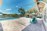 Located at 2801 NW 91st Ave in Coral Springs, Florida, this outdoor pool area offers a well-appointed recreational retreat designed for relaxation and leisure under the South Florida sun. The centerpiece is a generously sized swimming pool with crystal-clear blue water, accessible via stainless steel entry rails and steps with tiled depth markings. The pool features a distinctive L-shaped design, providing ample room for both swimming and wading.

Surrounding the pool is an expansive paver deck finished in a warm, neutral brick pattern that extends broadly in multiple directions, offering plentiful space for lounging and socializing. The deck is furnished with a variety of seating options, including Adirondack-style chairs in vibrant teal, sling-style lounge chairs, and shaded seating areas with colorful market umbrellas in teal and blue tones.

Lush tropical landscaping frames the entire area, with tall, slender palm trees providing natural shade and a distinctly Florida aesthetic. Mature trees along the perimeter add greenery and privacy. A Mediterranean-inspired clubhouse structure with a terracotta tile roof and warm stucco exterior is positioned poolside, adding architectural character to the space. The pool area is enclosed by a black iron fence, ensuring security while preserving open sightlines to the surrounding landscape and open sky beyond.