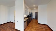 This single-floor unit offers a clean, modern aesthetic defined by rich dark hardwood-style flooring that runs throughout the entire space, creating a cohesive and warm foundation across every room. The open layout is anchored by a spacious living area bathed in natural light from two large windows that frame views of the surrounding urban skyline. A wall-mounted HVAC unit ensures year-round climate comfort directly within the living space.

The kitchen is efficiently designed with a galley-style layout, featuring dark lower cabinetry contrasted by white upper cabinets with open shelving, dark countertops with an undermount sink, and a full suite of stainless steel appliances including a refrigerator, stove, microwave, and dishwasher. Recessed lighting overhead adds a polished finish to the cooking area, which flows seamlessly toward the unit's entry door.

The bathroom is well-appointed, offering a bathtub, toilet, and vanity with a light countertop set against dark cabinetry. The hardwood-style flooring continues into this space, maintaining design continuity. Adjacent to the bathroom is a dedicated walk-in closet with built-in wire shelving, providing generous storage. The overall ambiance is bright, clean, and contemporary, with crisp white walls and a straightforward, functional floor plan.