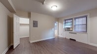 This single-floor unit is thoughtfully laid out to maximize its compact footprint. The main living area features rich, dark hardwood-style flooring that extends throughout, neutral beige walls, and a flush ceiling light fixture. A window fitted with blinds and a wall-mounted air conditioning unit provides both natural light and climate control, while a radiator beneath offers supplemental heat. An electrical panel is conveniently accessible on the main wall.

The bedroom is a well-proportioned space sharing the same hardwood-style flooring, with an open closet rod system for storage and a window that allows natural light to filter in. The room connects directly to the rest of the unit via a straightforward layout visible in the 3D floorplan.

The galley kitchen is lined with warm oak cabinetry above and below a continuous countertop, providing ample storage and prep space. A stainless steel sink, white refrigerator, and light tile flooring complete the functional design.

The bathroom showcases classic detailing, including white subway-style wall tiles accented by a black border, a black-and-white diamond-patterned floor, a full soaking tub with an overhead shower, and a white vanity with undermount sink. The overall ambiance is clean and practical throughout.