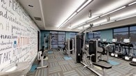 This modern fitness center is designed to provide a comprehensive and motivating workout environment. The space is anchored by a bold teal color scheme on the accent walls, complemented by a large, eye-catching mural featuring motivational phrases and typography that energizes the atmosphere. Bright, linear fluorescent lighting runs across the white tray ceiling, ensuring the entire floor is well-illuminated throughout the day. Large floor-to-ceiling windows line one side of the room, flooding the space with natural light and offering views of the surrounding urban landscape. The flooring consists of a multi-toned, striped carpet with accents of teal, gray, and beige, adding texture and visual interest underfoot. The fitness center is outfitted with a robust selection of commercial-grade equipment, including cable and weight machines, resistance training stations, and a row of cardio machines such as ellipticals and stationary bikes. The equipment is thoughtfully arranged to allow ample circulation and comfortable use throughout the space. Overall, the fitness center strikes a balance between functionality and contemporary design, creating an inviting and well-organized facility suited for a full range of exercise routines.