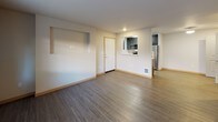 Located at 1060 Insignia Loop in Bremerton, Washington, this single-floor unit offers a clean, modern design throughout. The open living area is finished with durable wood-look vinyl flooring in a warm gray tone, recessed lighting, and neutral white walls accented by light wood baseboards. A pass-through window connects the living space directly to the kitchen, allowing for an open feel between the two areas. A secondary living or dining area features a glass-panel exterior door with a sidelight window, allowing natural light to flow in. The galley-style kitchen is well-appointed with dark charcoal cabinetry, white countertops, a stainless steel sink, and a full suite of stainless steel appliances including a refrigerator, range, microwave, and dishwasher. The flooring continues the wood-look vinyl theme for easy maintenance. The bedroom is carpeted for comfort and includes a window that brings in natural light, a ceiling-mounted light fixture, and ample wall space. The bathroom is efficiently designed with a full soaking tub and shower combo, a dark-cabinetry vanity with a light countertop, a large mirror, and wood-look flooring. In-unit laundry hookups are also present, adding convenience. The overall aesthetic is contemporary and functional.