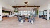 This expansive single-floor clubhouse spans a generously sized open-plan layout designed to serve as a vibrant communal gathering space. Light-toned hardwood flooring runs throughout, complementing the bright white walls and exposed wood ceiling beams that add architectural warmth. Soaring ceiling heights in select areas, particularly near the floor-to-ceiling windows and sliding glass doors, flood the interior with natural light while offering views of the surrounding outdoor landscape.

The main lounge area is anchored by a striking multi-bulb brass pendant chandelier and furnished with bold patterned curved sofas and natural wood stump side tables, creating an inviting, resort-style atmosphere. Adjacent seating zones offer additional neutral-toned sofas positioned around a wall-mounted display flanked by living moss panels—a recurring biophilic design element found throughout the space.

The fully equipped kitchen features stainless steel appliances, double wall ovens, dark wood cabinetry, a dramatic marble-topped island, and an eye-catching cityscape moss mural spanning the full length of the wall above the countertops. Decorative organic wall art accents add texture and personality.

A dedicated reception or leasing area near the entrance includes statement furniture, curated artwork, and additional moss wall installations. A separate enclosed room with glass partition walls appears to function as a private meeting or co-working space, rounding out this well-appointed communal facility.