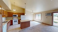 This single-floor unit offers a well-organized layout with distinct living, kitchen, and sleeping areas. The kitchen is a functional centerpiece, featuring warm medium-toned wood cabinetry extending to the upper walls, light countertops, and white appliances including a refrigerator, electric range, microwave, and dishwasher. A center island with a neutral countertop provides additional workspace and serves as a natural divider between the kitchen and the adjoining living and dining area. Pendant lighting illuminates both the island and kitchen, adding a decorative touch.

The open living and dining area transitions from tile flooring in the kitchen to plush carpet, creating a comfortable, defined space. Vaulted ceilings add a sense of openness, while large windows and a sliding glass door allow generous natural light and outdoor views. A ceiling fan and baseboard heating units are present throughout.

The bedroom is generously sized with carpeted flooring, a ceiling fan, windows for natural light, and a built-in shelving unit recessed into the wall. The bathroom includes tile flooring, a wide vanity with a white countertop, wood cabinetry below, and a large mirror above the sink.