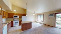This single-floor unit offers a well-organized layout with distinct living, kitchen, and sleeping areas. The kitchen is a functional centerpiece, featuring warm medium-toned wood cabinetry extending to the upper walls, light countertops, and white appliances including a refrigerator, electric range, microwave, and dishwasher. A center island with a neutral countertop provides additional workspace and serves as a natural divider between the kitchen and the adjoining living and dining area. Pendant lighting illuminates both the island and kitchen, adding a decorative touch.

The open living and dining area transitions from tile flooring in the kitchen to plush carpet, creating a comfortable, defined space. Vaulted ceilings add a sense of openness, while large windows and a sliding glass door allow generous natural light and outdoor views. A ceiling fan and baseboard heating units are present throughout.

The bedroom is generously sized with carpeted flooring, a ceiling fan, windows for natural light, and a built-in shelving unit recessed into the wall. The bathroom includes tile flooring, a wide vanity with a white countertop, wood cabinetry below, and a large mirror above the sink.