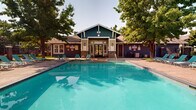 This outdoor amenity space centers around a generously sized swimming pool with clear, turquoise water, designed for both leisure and lap swimming. The pool is enclosed within a secure wrought iron fence, providing both safety and an open, airy feel. A wide, paved sun deck surrounds the pool on multiple sides and is furnished with numerous teal lounge chairs, offering ample space for sunbathing and relaxation. Mature deciduous trees are positioned throughout the area, providing natural shade and a lush, park-like ambiance. At the head of the pool, a craftsman-style building serves as the focal point of the space, featuring a steeply pitched gable roof, board-and-batten siding in a deep teal-blue hue with cream trim, stone veneer accents, and classic wall-mounted lantern fixtures flanking a set of glass-paneled double doors. The building's covered entry porch adds architectural character and provides a sheltered transition point. Additional seating areas with small tables are positioned near the building's entrance. Ornamental grasses and seasonal plantings accent the perimeter fencing, adding color and texture to the landscaped surroundings. A basketball hoop is visible in the background, further enhancing the recreational offerings of this well-maintained outdoor community space.