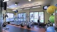 This fitness center is designed to support a well-rounded cardio workout routine. The space is outfitted with a row of treadmills, elliptical trainers, and a recumbent stationary bike, each equipped with individual screens for entertainment during exercise. The flooring features durable rubber matting in a warm brown tone, providing both safety and comfort underfoot. Overhead, a combination of pendant-style rectangular light fixtures and a ceiling fan ensures the space remains well-lit and well-ventilated throughout. Wall-mounted flat-screen televisions are positioned at either end of the room for easy viewing from multiple machines. Large windows with horizontal blinds line the walls, allowing natural light to fill the space while offering views of the surrounding greenery outdoors. The walls are finished in a neutral olive-green tone, contributing to a calm and focused workout environment. The overall layout is clean and organized, with equipment arranged in a single, open row that maximizes accessibility and allows for easy movement throughout the room.