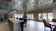 This elegantly appointed single-floor space seamlessly blends classic architectural details with contemporary furnishings to create a refined yet relaxed atmosphere. The most striking feature is the ornate pressed-tin ceiling, finished in a polished silver tone and adorned with intricate repeating medallion patterns — a design element that immediately draws the eye upward and lends the room a distinctly sophisticated character. Recessed lighting embedded within the ceiling tiles, alongside a sleek modern chandelier, ensures the space is well-lit at any time of day.

Large, evenly spaced windows line the walls, flooding the interior with generous natural light and offering pleasant views of the surrounding greenery and landscaping. Light-toned, large-format tile flooring reflects this natural light beautifully, enhancing the sense of openness throughout the room. Wainscoting and paneled wall detailing add further architectural depth and refinement.

The room is thoughtfully furnished for leisure and socializing. A full-size billiards table takes center stage, while a tall bar-height counter accompanied by black leather bar stools provides a casual seating area ideal for gathering. The overall ambiance is one of upscale recreation — a space designed equally for relaxed entertaining and active enjoyment.