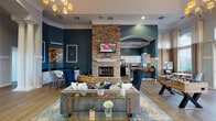This thoughtfully designed single-story community space blends sophisticated style with versatile, amenity-rich functionality. The entry foyer welcomes visitors with warm hardwood-style flooring, decorative columns, arched architectural details, and an elegant chandelier overhead, setting the tone for the refined interiors beyond.

The expansive main lounge area serves as the heart of the space, anchored by a dramatic floor-to-ceiling stacked stone fireplace with a mounted television above. Bold teal accent walls contrast beautifully with white crown molding and trim throughout, creating a polished yet inviting atmosphere. Comfortable seating arrangements, including plush sofas and wingback chairs centered around a glass-and-gold coffee table, encourage relaxed socializing.

An open-plan kitchen features white cabinetry, dark granite countertops, black appliances, and a breakfast bar with leather-upholstered bar stools—ideal for entertaining. Adjacent areas include a dedicated gaming nook with an arcade table and a foosball table, adding a recreational dimension to the space.

A sunlit seating area with soaring arched windows and French doors opens directly to an outdoor pool area, flooding the interior with natural light and offering refreshing views of the surrounding landscaping.