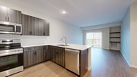 Located at 1130 Hazeltine Blvd in Chaska, MN, this thoughtfully designed 2-bedroom, 1-bathroom unit offers a modern, open-concept floor plan across a single level. The kitchen serves as a central highlight, featuring dark wood-grain cabinetry, a classic white subway tile backsplash, quartz countertops, a peninsula with an undermount double sink, and a full suite of stainless steel appliances including a refrigerator, range, microwave, and dishwasher. The kitchen flows seamlessly into the living room, which is finished with durable wood-style plank flooring, built-in shelving for display or storage, and a sliding glass door that opens to a private balcony with outdoor views. Both bedrooms are carpeted and receive natural light through large windows with blinds. The primary bedroom includes a closet and generous floor space. The shared bathroom is well-appointed with a wood-tone vanity, white quartz countertop, matte black hardware and fixtures, a soaking tub, and wood-look tile flooring. A dedicated in-unit laundry area houses a full-size front-loading washer and dryer with wire shelving overhead, adding functional convenience to this well-rounded home.