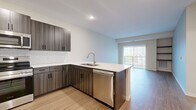 Located at 1130 Hazeltine Blvd in Chaska, MN, this thoughtfully designed 2-bedroom, 1-bathroom unit offers a modern, open-concept floor plan across a single level. The kitchen serves as a central highlight, featuring dark wood-grain cabinetry, a classic white subway tile backsplash, quartz countertops, a peninsula with an undermount double sink, and a full suite of stainless steel appliances including a refrigerator, range, microwave, and dishwasher. The kitchen flows seamlessly into the living room, which is finished with durable wood-style plank flooring, built-in shelving for display or storage, and a sliding glass door that opens to a private balcony with outdoor views. Both bedrooms are carpeted and receive natural light through large windows with blinds. The primary bedroom includes a closet and generous floor space. The shared bathroom is well-appointed with a wood-tone vanity, white quartz countertop, matte black hardware and fixtures, a soaking tub, and wood-look tile flooring. A dedicated in-unit laundry area houses a full-size front-loading washer and dryer with wire shelving overhead, adding functional convenience to this well-rounded home.