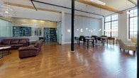 The WiFi Lounge and Library is a generously proportioned communal space that seamlessly blends industrial character with refined comfort. Soaring ceilings with exposed wooden beams and industrial-style track lighting give the space an open, loft-like quality, while large multi-pane windows flood the interior with natural light and offer views of the surrounding cityscape.

Rich hardwood flooring runs throughout, tying together several distinct functional zones. A casual lounge area features striped upholstered armchairs arranged around a small coffee table, ideal for relaxed conversations or informal work sessions. A separate seating nook, partially enclosed by glass-panel partitions, includes leather sofas and is anchored by a striking stone-clad fireplace with a mounted flat-screen TV above it — creating an inviting retreat-like atmosphere.

The library section is clearly defined, with teal-painted built-in bookshelves stocked with reading material. Adjacent to this, a dedicated work area offers long communal tables with pendant lighting overhead, positioned against a textured stone-look feature wall. Glass-walled rooms along the perimeter provide semi-private spaces for focused work, each equipped with bar-height seating and additional workspace, ensuring the lounge accommodates a wide range of uses throughout the day.