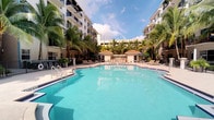 This outdoor amenity area centers around a generously sized resort-style swimming pool with striking turquoise water, offering a striking focal point within the complex. The pool's curvilinear design adds a relaxed, leisure-oriented aesthetic, complemented by clean white pool decking that surrounds the water on multiple sides. A dedicated cabana structure with a tiled roof sits at the far end of the pool area, providing shaded seating and a gathering space for residents or guests. Rows of lounge chairs are arranged along the pool's edge, ideal for sunbathing or relaxing poolside. Tall, mature palm trees are thoughtfully integrated throughout the space, enhancing the tropical ambiance and offering natural shade. The pool is flanked on both sides by a modern multi-story building featuring balconies that overlook the amenity area, creating an enclosed courtyard-like setting. Umbrella-shaded seating areas are also available for additional comfort. The overall atmosphere is bright, open, and resort-inspired, with abundant natural light and a well-maintained landscape contributing to a vibrant, sun-soaked outdoor environment suited for warm-weather living.