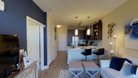 This single-floor residence offers a well-designed open-concept layout that seamlessly connects the kitchen, dining, and living areas into one cohesive space. The kitchen is a standout feature, boasting dark espresso cabinetry with open shelving, a stainless steel refrigerator, a tiled backsplash, and light-toned countertops. A peninsula-style breakfast bar with sleek black bar stools provides both additional prep space and casual seating, while pendant lighting adds a warm, modern touch overhead. Track lighting further enhances the kitchen's functionality and ambiance. The living area flows naturally from the kitchen and features light wood-style flooring that runs throughout, contributing to a bright and airy feel. A bold navy blue accent wall creates a striking visual anchor near the entryway, adding depth and personality to the otherwise soft, neutral color palette. The entry corridor provides a sense of arrival, with clean white doors and easy access to adjacent rooms. The overall aesthetic is contemporary and inviting, combining practical design elements with stylish details that make the space feel both livable and refined.