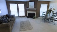 This single-story living room is defined by its warm, earthy aesthetic, with deep brown walls that create an inviting and grounded atmosphere. The focal point of the room is a white-tiled wood-burning fireplace, topped with a wooden mantel that displays candles and a large framed mirror, adding depth and elegance to the space. Vaulted ceilings lend a sense of openness and airiness, enhancing the room's overall spaciousness. Soft, neutral wall-to-wall carpeting runs throughout, complemented by a plush area rug positioned in the center of the room. Multiple windows fitted with white horizontal blinds allow natural light to filter in from several directions, brightening the interior while maintaining privacy. A small decorative plant near the fireplace hearth adds a subtle touch of greenery. Additional seating is visible toward the right side of the room, suggesting a versatile layout suitable for both relaxing and entertaining. The overall ambiance is casual yet comfortable, combining functional design elements with warm, neutral tones for a cohesive and welcoming living environment.