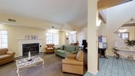 This single-floor space offers a warm and functional environment designed for comfort and community engagement. The main lounge area is anchored by a classic marble-surround fireplace, which serves as a natural focal point and lends the room a cozy, inviting ambiance. Soft, neutral yellow walls and carpeted flooring contribute to the space's warm atmosphere throughout.

The seating area is generously appointed with a sage green sofa and tan leather armchairs arranged around a decorative coffee table, creating an ideal setting for relaxed conversation or leisure. Large multi-pane windows allow ample natural light to filter into the room, brightening the interior and offering views of the surrounding outdoors.

Adjacent to the lounge, a dining or activity area is visible, furnished with a wooden table and chairs, providing additional functional space for various uses. A dedicated workstation or bulletin board area is positioned near the entrance, offering practical resources for occupants, with tiled flooring distinguishing it from the carpeted lounge. Recessed and wall-mounted lighting throughout ensures the space remains well-lit during evening hours. The overall layout is open and accessible, combining comfort with practicality in a thoughtfully arranged single-level setting.