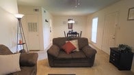 This single-floor residence offers a welcoming and functional layout designed for comfortable everyday living. The living area serves as the heart of the home, featuring plush carpeted flooring in a neutral tone that adds warmth underfoot. A large, dark upholstered loveseat anchors the seating area, accented with decorative throw pillows in rich earthy tones. The textured ceiling adds subtle character to the room, while a pendant light fitting provides a central focal point overhead.

The living space flows seamlessly into a dining area, which is highlighted by a classic chandelier that casts a soft, inviting glow over a dark wood dining table and chairs. Decorative wall art frames the dining nook, adding a personal touch to the space. A large window on the far wall allows natural light to filter into the room, brightening the open-plan area throughout the day.

Adjacent to the living space, closet and utility access is conveniently integrated into the layout, contributing to the home's practical design. The overall ambiance of this residence is warm and understated, with a neutral color palette of soft beiges and whites that creates a calm, livable atmosphere well-suited for both relaxing and entertaining.
