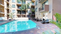 This inviting outdoor amenity space is centered around a generously sized swimming pool with crystal-clear turquoise water, framed by a classic terracotta-tiled sundeck that evokes a warm, resort-like atmosphere. The courtyard is nestled within a multi-story Mediterranean-inspired building, whose warm golden stucco façade, ornamental balconies, and columned detailing create an elegant architectural backdrop on all sides.

The pool deck is well-appointed with multiple sleek dark-framed chaise lounges and ottomans fitted with plush cream cushions, providing ample space for relaxation in the sun. A shaded outdoor dining area with a market umbrella, table, and chairs offers an additional gathering spot for residents. Lush tropical landscaping, including tall swaying palms, sago palms, and a variety of tropical foliage, adds vibrant greenery throughout the courtyard, enhancing the resort ambiance.

Glass-paned doors connect the outdoor deck to an interior lounge area, where wicker sofas and chaise lounges with neutral cushions provide a comfortable transitional space between indoors and out. The tile-paved pathways wind through layered plantings, creating charming garden vignettes. Abundant natural sunlight floods the open-air courtyard, making this a bright and welcoming communal retreat throughout the day.
