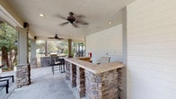This outdoor amenity space is thoughtfully designed to provide a comprehensive recreational experience. At the heart of the area is a generously sized, covered pavilion featuring a built-in bar constructed with stacked stone bases and a tiled countertop, ideal for outdoor entertaining. Ceiling fans are mounted overhead to ensure comfort during warm weather, while recessed lighting and shiplap-accented walls add a polished, finished aesthetic to the covered area. Additional built-in grilling and prep stations extend the outdoor kitchen functionality.

The centerpiece of the complex is a resort-style swimming pool with a curved, free-form design framed by a broad brick-paved deck. Cushioned lounge chairs are positioned poolside, offering ample space for relaxation. Umbrella-shaded tables and seating clusters provide comfortable gathering spots throughout the deck area.

The surrounding courtyard is finished with decorative brick pavers and is framed by mature trees and well-maintained landscaping beds. The multi-story brick exterior buildings, featuring bay windows, balconies, and pitched rooflines, create an attractive and cohesive architectural backdrop. Walkways meander through the greenery, connecting all areas of the community with a welcoming, park-like atmosphere.
