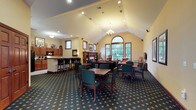 This single-story gathering space combines comfort and functionality in a warm, welcoming environment. The focal point of the room is its dramatic vaulted ceiling, which rises to a prominent peak and is highlighted by a classic chandelier and recessed lighting that together create a bright, airy atmosphere. A large arched window flanked by additional rectangular windows along the far wall floods the space with natural light and provides views of lush greenery outside.

The layout is thoughtfully divided into distinct areas. A dining section near the center features multiple dark wood tables with green upholstered chairs, arranged to accommodate groups of various sizes. Toward the back, a cozy lounge area with sofas and armchairs surrounds the fireplace, providing a more relaxed setting for conversation or leisure. A decorative mantel adds architectural character to this corner of the room.

Along one wall, a kitchenette with dark cabinetry, a countertop bar, and bar stools offers a casual service area. The patterned, dark-toned carpeting anchors the space and complements the warm yellow walls and wood trim throughout, contributing to an overall ambiance that is inviting and well-appointed.