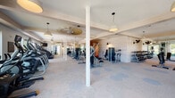 This expansive fitness center is thoughtfully designed to accommodate a wide range of workout styles across multiple interconnected zones. The layout transitions between warm hardwood flooring in open stretching and functional training areas and low-pile carpet tiles in the strength and cardio sections, providing both aesthetic variety and practical surface options for different exercises.

The strength training area is well-stocked with a comprehensive selection of cable machines, a power rack with bench, leg press, and a full dumbbell rack, ensuring users have access to a thorough range of resistance equipment. Large floor-to-ceiling windows line several sections of the facility, flooding the space with natural light and offering views of the surrounding greenery. Wall-mounted mirrors further enhance the sense of openness while serving a functional purpose for form monitoring.

The cardio zone features a row of treadmills and stationary bikes, positioned near the windows to make workouts more engaging. Wall-mounted televisions are strategically placed for convenient viewing. Architectural details including white-painted structural columns, exposed beam ceilings, and light brick accent walls lend the space a refined, modern aesthetic. Pendant lighting and ceiling fans complete the design. A dual water fountain and bottle-filling station, set against a textured brick wall, adds a practical and polished amenity to the facility.