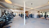 This expansive fitness center is thoughtfully designed to accommodate a wide range of workout styles across multiple interconnected zones. The layout transitions between warm hardwood flooring in open stretching and functional training areas and low-pile carpet tiles in the strength and cardio sections, providing both aesthetic variety and practical surface options for different exercises.

The strength training area is well-stocked with a comprehensive selection of cable machines, a power rack with bench, leg press, and a full dumbbell rack, ensuring users have access to a thorough range of resistance equipment. Large floor-to-ceiling windows line several sections of the facility, flooding the space with natural light and offering views of the surrounding greenery. Wall-mounted mirrors further enhance the sense of openness while serving a functional purpose for form monitoring.

The cardio zone features a row of treadmills and stationary bikes, positioned near the windows to make workouts more engaging. Wall-mounted televisions are strategically placed for convenient viewing. Architectural details including white-painted structural columns, exposed beam ceilings, and light brick accent walls lend the space a refined, modern aesthetic. Pendant lighting and ceiling fans complete the design. A dual water fountain and bottle-filling station, set against a textured brick wall, adds a practical and polished amenity to the facility.