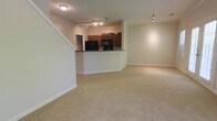 This two-story residence offers a thoughtfully designed layout that maximizes both functionality and comfort. The main floor showcases an open-concept living and kitchen area, creating a cohesive and inviting space ideal for everyday living and entertaining. The living area is generously sized, featuring wall-to-wall neutral carpeting that adds warmth underfoot and a clean, consistent aesthetic throughout. A staircase with white-painted trim leads to the upper level, adding a classic architectural touch to the interior.

The kitchen is positioned at the heart of the main floor and features rich, dark wood cabinetry paired with dark countertops, creating a striking contrast against the light-toned walls. Black appliances, including a refrigerator and range, lend a modern and uniform look to the cooking space. A raised breakfast bar serves as a natural divider between the kitchen and living area, providing additional seating and prep space while maintaining the open feel of the floor plan.

Track lighting runs along the ceiling in both the kitchen and the adjoining living space, ensuring ample, adjustable illumination throughout. The walls are finished in a soft, neutral tone that enhances the brightness of the interior. The overall ambiance is clean, functional, and move-in ready, offering a comfortable foundation for a variety of living arrangements across both floors.
