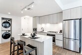 This contemporary single-floor residence is designed with a clean, modern aesthetic throughout. The open-concept kitchen is a standout feature, showcasing light wood cabinetry extending to the ceiling, stainless steel appliances including a gas range, built-in microwave, and refrigerator, all centered around a sleek white quartz island with bar seating. Warm hardwood flooring runs throughout the main living areas, connecting the kitchen seamlessly to the living room, which is bathed in natural light through large, black-framed windows offering urban views. The living space is generous, accommodating multiple seating arrangements with ease.

The bedrooms are well-proportioned, each featuring large windows, hardwood or wood-tone flooring, and ample closet space. Two bathrooms serve the unit: one features dark hexagonal floor tile, a light wood floating vanity, and black fixtures, while the primary bathroom offers a double-sink vanity, blue-grey hexagonal tile flooring, and a separate shower with subway tile surround. A stacked washer and dryer unit is conveniently located within the residence. The overall ambiance is bright, minimalist, and thoughtfully appointed throughout.