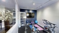 This dedicated fitness studio is thoughtfully designed to provide an effective and comfortable workout environment. The space features white crown molding and light gray walls that create a clean, bright atmosphere, with a bold navy blue accent wall serving as a striking focal point. A large wall-mounted flat-screen TV is positioned on the accent wall, making it ideal for following workout videos or streaming fitness classes. Track lighting runs along the ceiling, ensuring the space is well-lit throughout. The studio is equipped with multiple stationary cycling bikes, offering a spin-class-style experience, along with exercise mats and stability balls for stretching, yoga, or floor-based workouts. Rubber flooring throughout provides both safety and durability for high-intensity training. A glass-paned French door connects the studio to an adjacent space, adding a sense of openness while maintaining a distinct workout zone. Shelving along the accent wall provides convenient storage for equipment and accessories. The layout is efficient and uncluttered, making it easy to move between different exercise stations. Overall, this fitness studio combines functionality with a polished aesthetic, delivering a professional-grade training environment.