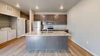 This thoughtfully designed one-bedroom, one-bathroom unit offers a functional and modern living experience across a single floor. The open-concept kitchen serves as a central highlight, featuring rich dark wood cabinetry with sleek bar-pull hardware, granite countertops, a large kitchen island with an undermount sink, and a full suite of stainless steel appliances including a refrigerator, range, and microwave. Light wood-tone flooring flows throughout the main living areas, complementing the neutral gray color palette and recessed lighting overhead. A distinctive industrial touch is carried throughout the space via exposed metallic ductwork, adding character and visual interest to the ceilings. The living area is bright and airy, benefiting from multiple windows dressed with blinds and vertical shades that invite natural light. The bedroom is carpeted for comfort, with a window providing additional natural light. The bathroom includes a walk-in shower, a dark wood vanity with granite countertop, and light wood-tone flooring. Three closets provide ample storage, while a dedicated laundry room houses a full-size top-loading washer and dryer with overhead shelving — a practical and convenient amenity within the unit.