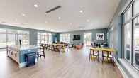 Located at 8710 SW 71st Ave in Ocala, Florida, this single-story community clubhouse is thoughtfully designed to serve as a welcoming social hub. The interior is defined by a cohesive color palette of soft teal-blue walls, crisp white trim, and light wood-look flooring that flows seamlessly throughout the space.

The main gathering area is generously proportioned, offering multiple seating configurations to accommodate a variety of activities. A lounge zone features coral upholstered chairs arranged around a sculptural coffee table atop a patterned area rug, anchored by a wall-mounted flat-screen television. Nearby, a bistro-style dining section provides long communal tables with mixed seating, ideal for group gatherings or casual meals.

Equestrian-themed artwork adorns the walls, adding a nod to Ocala's renowned horse country heritage. Large windows and multiple glass doors flood the interior with natural light and offer direct access to an outdoor patio, extending the usable space and providing open views of the surrounding landscape.

The fully equipped kitchen is a standout feature, showcasing white shaker cabinetry, a colorful tiled backsplash, stainless steel appliances including a wall oven and side-by-side refrigerator, a central island, and stylish pendant lighting. Recessed lighting throughout ensures the entire space remains bright and inviting at all hours.