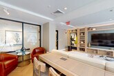 Located at 1440 Beacon St in Boston's Brookline neighborhood, this thoughtfully designed communal lounge offers a refined and inviting atmosphere. The single-floor space is finished with light herringbone hardwood flooring throughout, which lends warmth and visual interest to the room. Recessed lighting and a tray ceiling detail provide a clean, polished aesthetic overhead.

The room is anchored by a large, built-in shelving and media wall crafted from light natural wood, featuring display niches, decorative objects, a prominent large-screen TV, and an integrated lower media console. A vibrant abstract artwork adds a bold focal point within the shelving unit. A generously sized sectional sofa, accented with an array of decorative pillows, is centered on a colorful abstract area rug, creating a comfortable and visually dynamic seating area.

A pair of rich red leather armchairs is positioned near expansive floor-to-ceiling windows that frame views of Beacon Street and its tree-lined surroundings, flooding the space with natural light. Frosted glass sliding partition panels with metal frames divide the lounge from an adjacent area, offering flexibility in the room's layout. A long conference-style table with upholstered seating near the entrance provides a functional workspace within the lounge, making this space equally suited for social gatherings and productive collaboration.