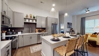 This single-floor residence is designed with a contemporary aesthetic, combining functional layouts with stylish finishes. The open-concept kitchen serves as a central hub, showcasing gray shaker-style cabinetry, dark tile backsplash, white quartz countertops, and stainless steel appliances. A spacious island with seating for multiple guests provides additional prep space and a casual dining option, illuminated by sleek pendant lighting overhead. Wood-style flooring flows throughout the main living areas, adding warmth to the modern palette.

The living room is bright and welcoming, benefiting from large windows and a glass door that opens to a private balcony, drawing in natural light. The primary bedroom features notably tall ceilings that enhance the sense of space, with an en-suite bathroom finished in white subway tile, a glass-enclosed walk-in shower, gray vanity cabinetry, and matte black fixtures. A second bedroom offers a comfortable layout with a large window and space for both sleeping and a dedicated workspace. The secondary bathroom includes a soaking tub and shower combination with white tile surround and coordinating matte black hardware throughout.