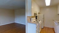 This single-floor residence offers a practical and functional layout that seamlessly connects the kitchen and living areas. The kitchen is equipped with white upper cabinetry that provides ample storage, a coil electric range, an over-the-range ventilation hood, and a full-size refrigerator — all finished in white for a clean, cohesive look. Light-toned laminate countertops complement the cabinetry, and the space is designed efficiently to maximize utility. A partial wall divides the kitchen from the adjoining living area, creating a sense of separation while maintaining an open feel. The living area features warm hardwood-style flooring that extends throughout, adding a welcoming and homey character to the space. Neutral-toned walls contribute to a bright and airy ambiance, while standard baseboards and subtle wall outlets reflect a straightforward, no-frills design aesthetic. The overall layout is compact yet well-organized, making efficient use of the available space. This single-level residence is well-suited for those seeking a low-maintenance living environment with all the essential features conveniently arranged on one floor.